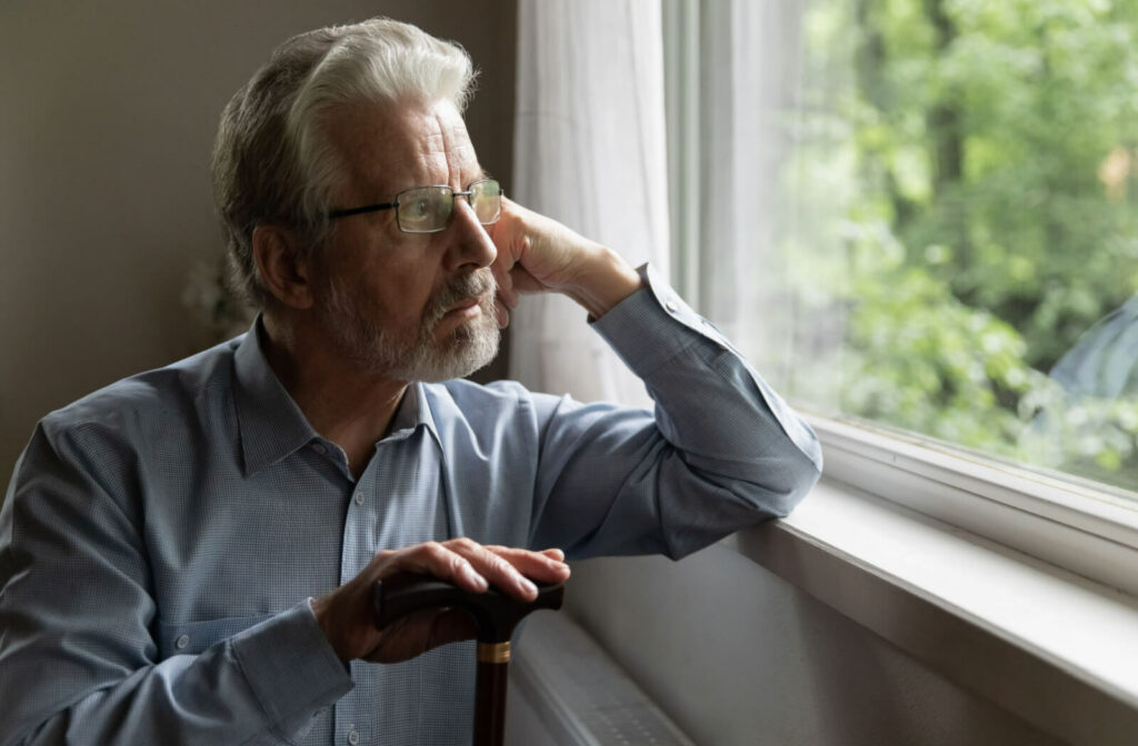 An older adult man with glasses leaning on a windowsill and resting his right hand on the top of a cane while looking out the window with a serious expression